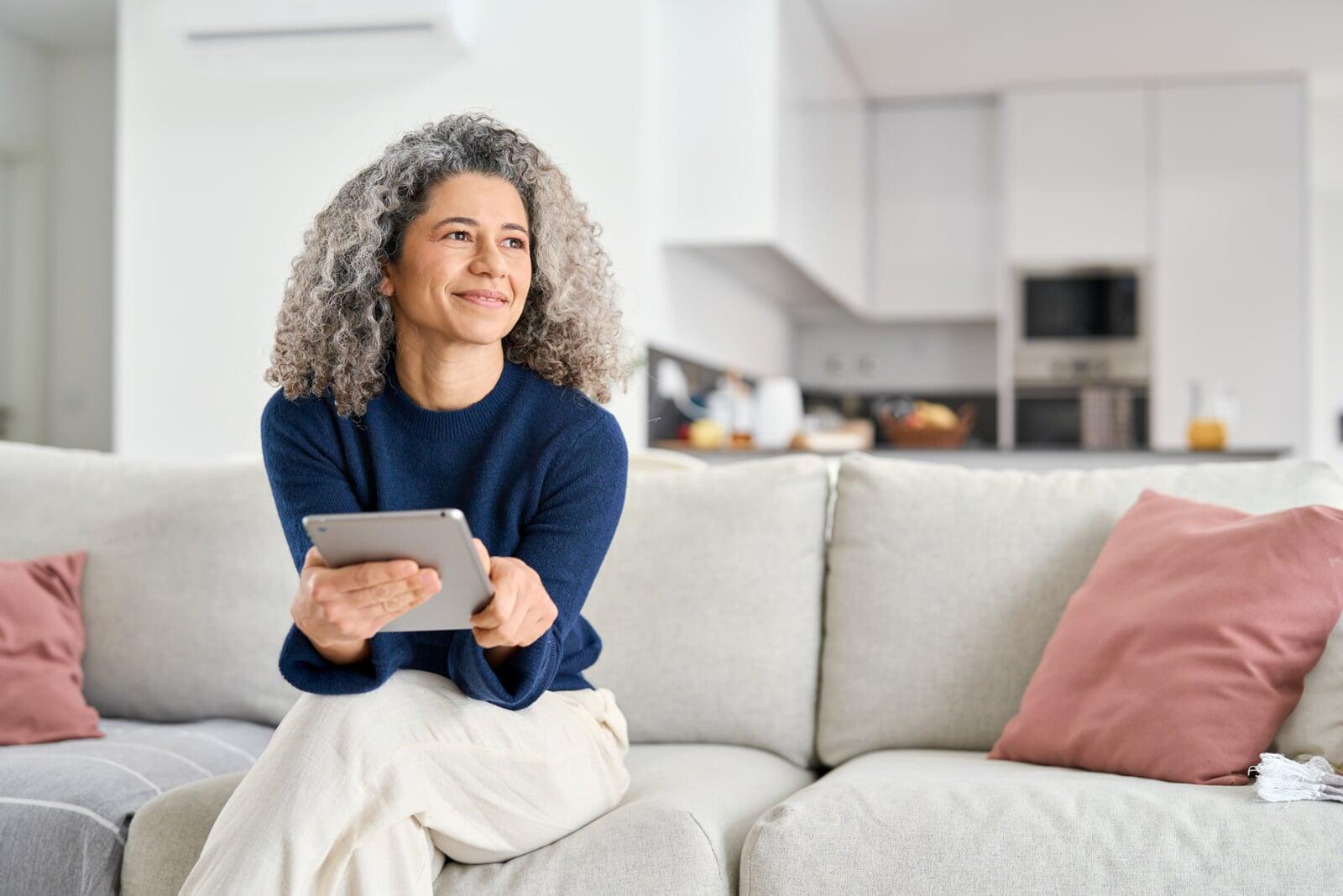 Schicke Frau mit grauen locken sitzt mit Tablet in der Hand auf der Couch.
