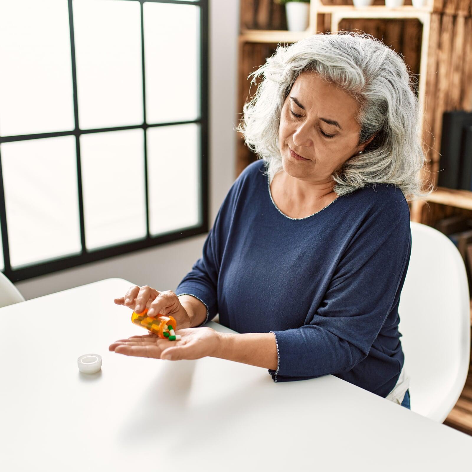 Frau mit grauen Haaren nimmt ihre Medikamente am Küchentisch ein.