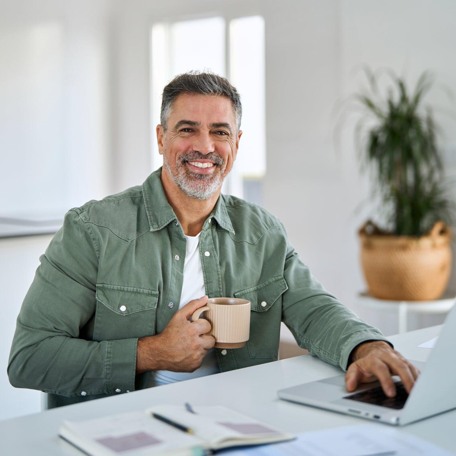Attraktiver älterer Herr sitzt mit Kaffeetasse und Laptop am Tisch und lächelt in die Kamera.