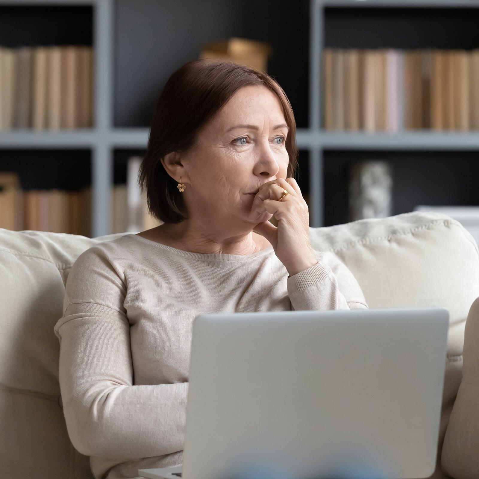 Grübelnde Frau sitzt mit ihrem Laptop auf der Couch.