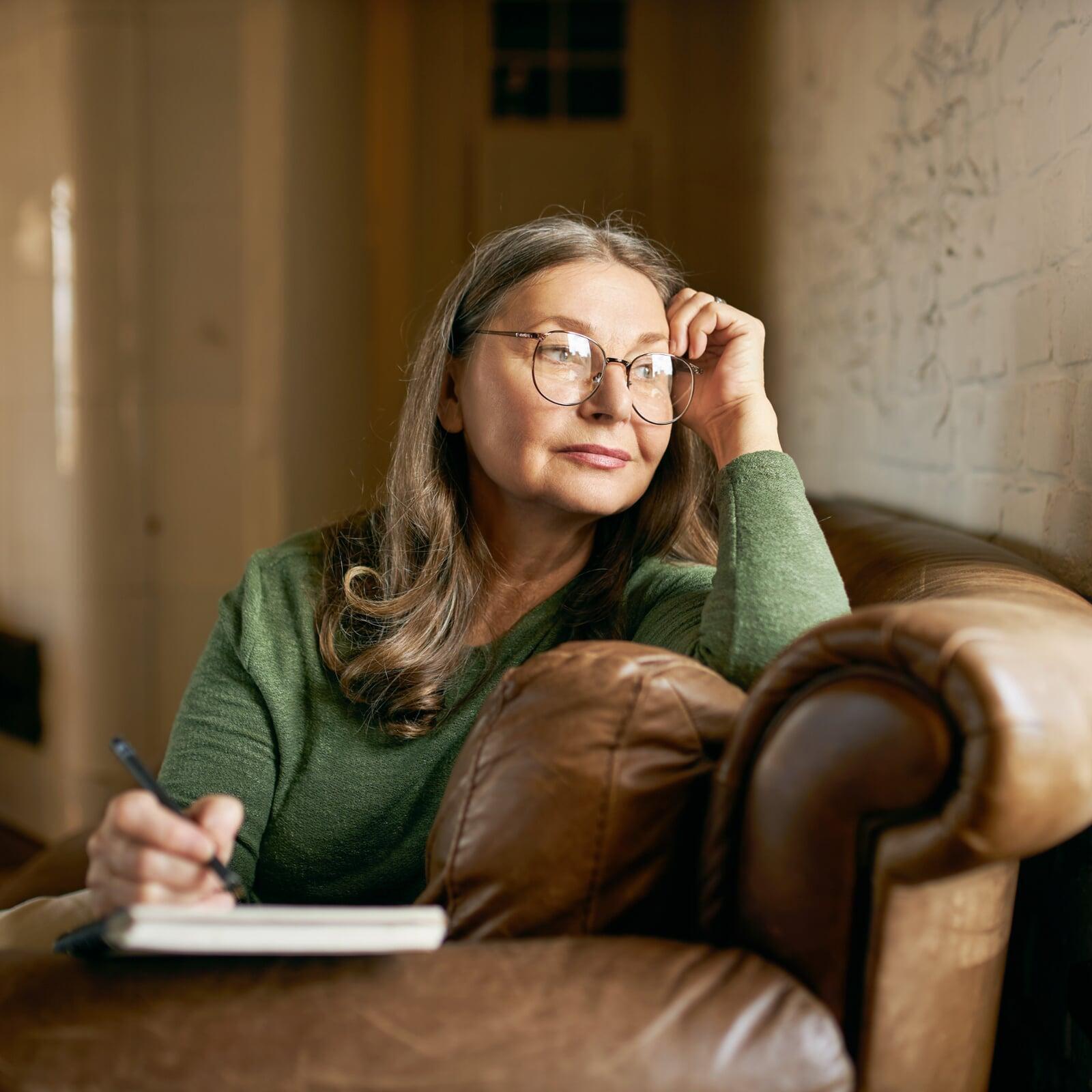 Frau in grünem Pullover mit Brille sitzt nachdenklich auf der Couch mit einem Stift in der rechten Hand, vor ihr ein Block Papier. 
