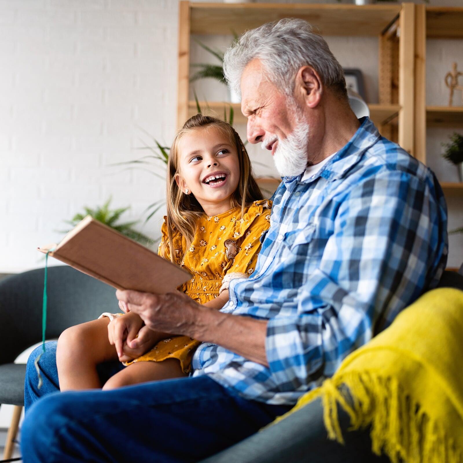 Älterer Herr mit weissem Vollbart liest lachendem Kind aus einem Buch vor.